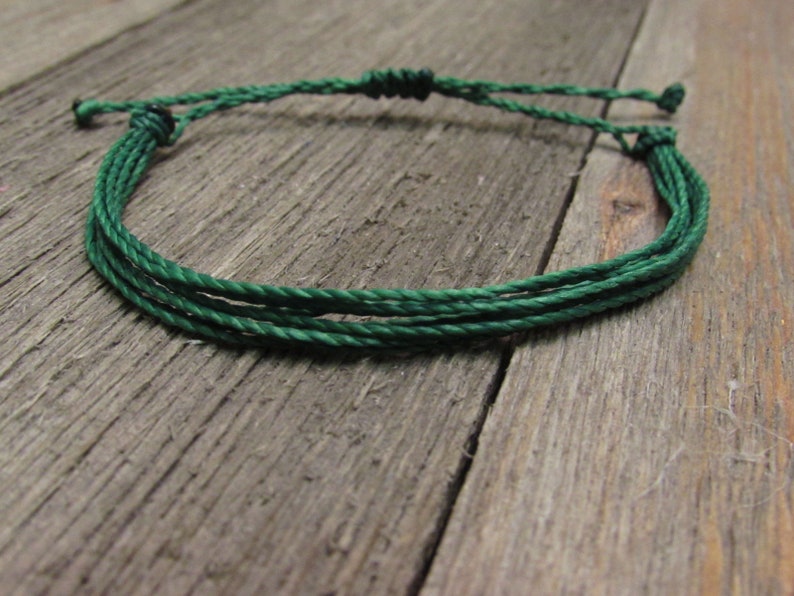 May include: A dark green adjustable bracelet made of multiple strands of waxed cord. The bracelet is knotted at the ends for adjustment and closure. The background is a weathered wooden surface.