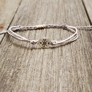 May include: A gray and white braided bracelet with a silver compass charm. The bracelet is tied with a knot on each end.