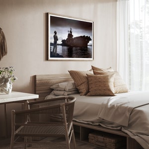 May include: A bedroom with a wooden bed frame, a white duvet, and a brown throw pillow. A framed black and white photograph of a ship hangs on the wall above the bed. A wooden desk with a white surface is in the foreground.