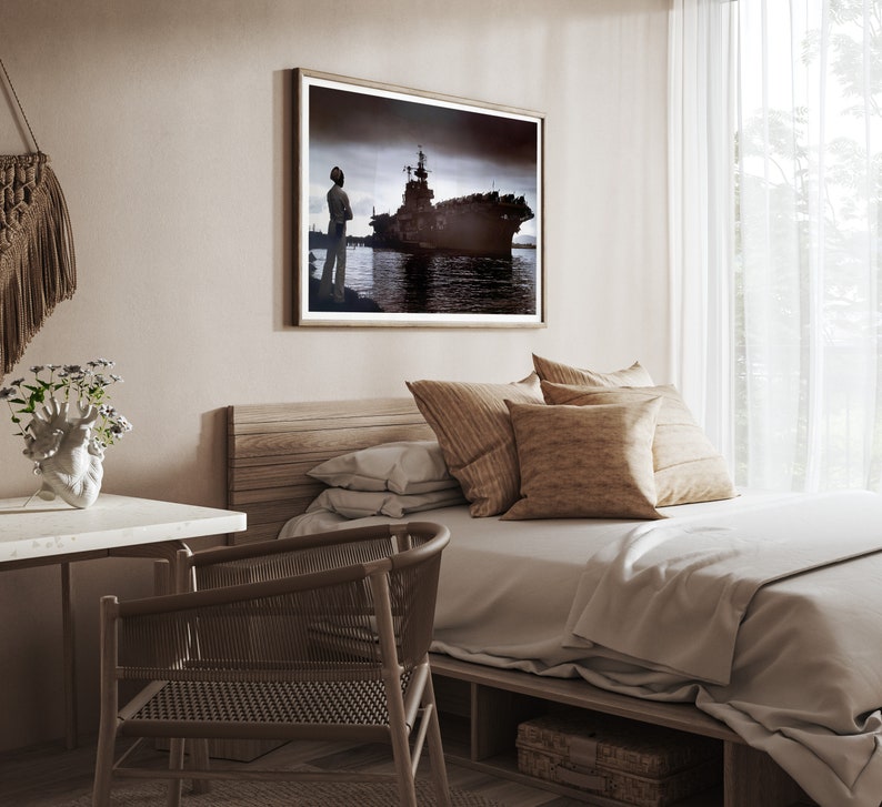 May include: A bedroom with a wooden bed frame, a white duvet, and a brown throw pillow. A framed black and white photograph of a ship hangs on the wall above the bed. A wooden desk with a white surface is in the foreground.