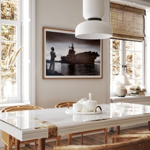 May include: A white dining table with a glass top and a white chair with a woven back. The table is set with a white teapot and cups. A framed photograph of a ship and a person standing on a dock is hanging on the wall behind the table.