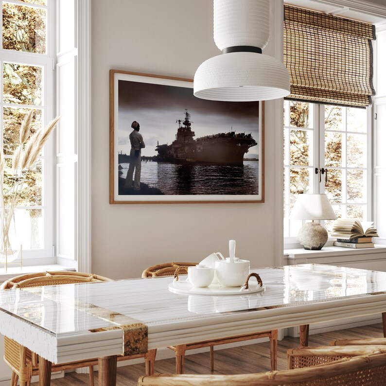 May include: A white dining table with a glass top and a white chair with a woven back. The table is set with a white teapot and cups. A framed photograph of a ship and a person standing on a dock is hanging on the wall behind the table.