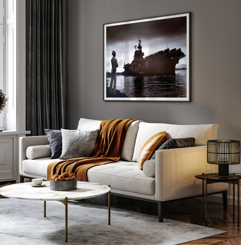 May include: A white sofa with a brown throw blanket and pillows, a black and gold side table with a lamp, and a coffee table with a bowl of oranges. A framed black and white photo of a ship hangs on the wall.