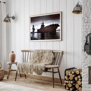 May include: A wooden bench with a faux fur throw blanket, a brown clay vase, and a framed black and white photograph of a ship in a harbor. The wall behind the bench is white wood paneling. There is a black metal fireplace with a wood log holder to the right of the bench.