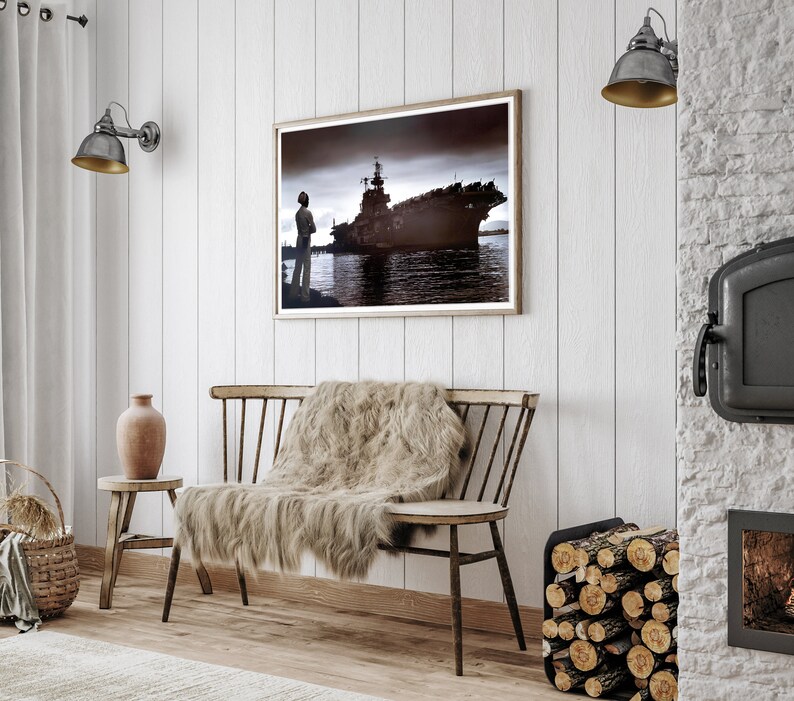May include: A wooden bench with a faux fur throw blanket, a brown clay vase, and a framed black and white photograph of a ship in a harbor. The wall behind the bench is white wood paneling. There is a black metal fireplace with a wood log holder to the right of the bench.