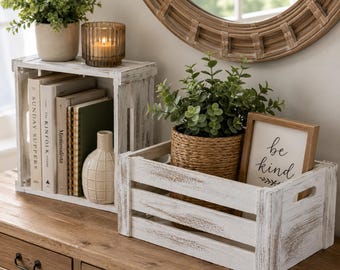 May include: Two white distressed wooden crates used for storage. The upright crate holds books, a small vase, and a green plant. The horizontal crate contains a woven basket with a green plant and a framed sign with the text be kind.