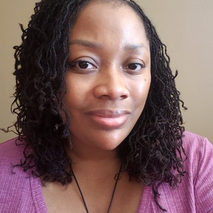 Peut inclure: Une femme avec des cheveux bruns fonc&eacute;s coiff&eacute;s en locs porte une chemise violette avec un collier noir et un pendentif.