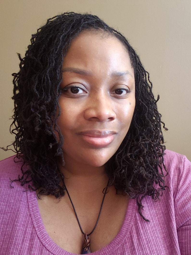 Peut inclure: Une femme avec des cheveux bruns fonc&eacute;s coiff&eacute;s en locs porte une chemise violette avec un collier noir et un pendentif.
