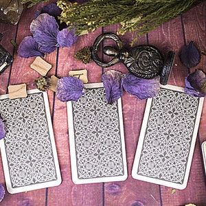 Peut inclure: Trois cartes de tarot avec des motifs noirs et blancs complexes sur une surface en bois violet. Les cartes sont entour&eacute;es de p&eacute;tales de fleurs s&eacute;ch&eacute;es violettes et d'une petite figurine en m&eacute;tal.