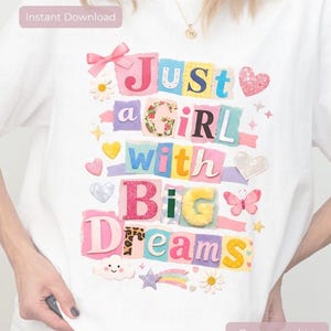 以下が含まれることがあります： 「Just a Girl with Big Dreams」と書かれたカラフルなグラフィックが特徴の白いTシャツ。ハート、花、蝶などの装飾要素がデザインされています。テキストは、さまざまなフォントとテクスチャを使用したコラージュスタイルです。