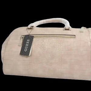 May include: A light pink Guess handbag with a textured, crocodile-like pattern. The bag has a top handle, a zipper closure, and a tag that reads "GUESS". The bag is set against a black background.