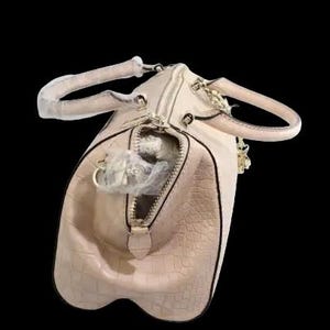 May include: A light pink, textured handbag with a zipper closure and two handles. The bag has a gold-colored chain and zipper pull. The interior of the bag is visible, with a white object inside. The background is black.