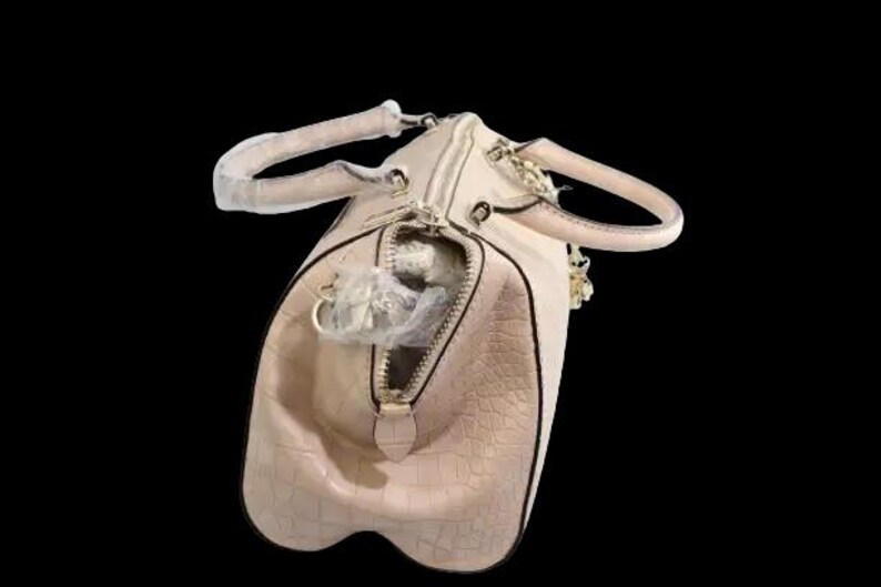 May include: A light pink, textured handbag with a zipper closure and two handles. The bag has a gold-colored chain and zipper pull. The interior of the bag is visible, with a white object inside. The background is black.