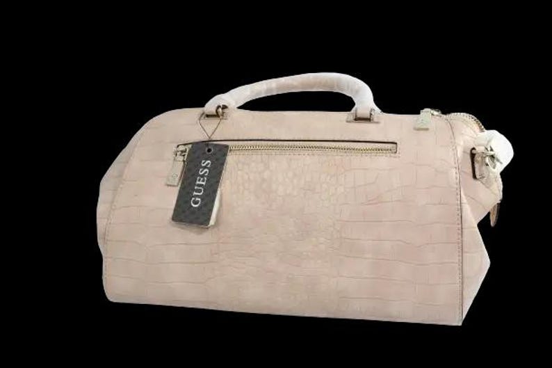 May include: A light pink Guess handbag with a textured, crocodile-like pattern. The bag has a top handle, a zipper closure, and a tag that says "GUESS". The bag is set against a black background.