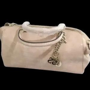 May include: A light beige handbag with a textured, crocodile-like pattern. The bag features a white handle, a silver chain with charms, and a zipper closure. The bag is set against a black background.