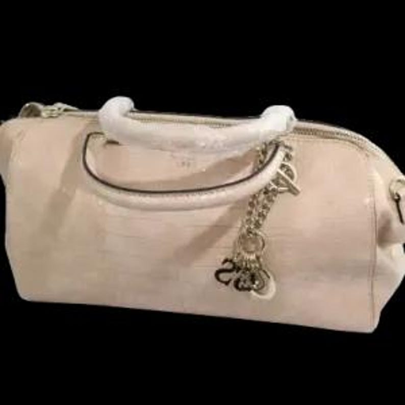 May include: A light beige handbag with a textured, crocodile-like pattern. The bag features a white handle, a silver chain with charms, and a zipper closure. The bag is set against a black background.