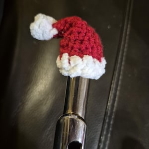 May include: A red and white crocheted Santa hat, designed to fit over the end of a silver flute. The hat is made of red yarn with a white fluffy trim and a white pom-pom. The flute is made of shiny metal.