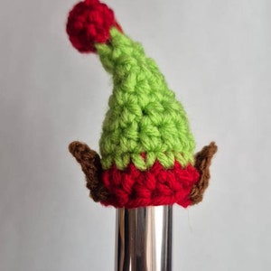 May include: A green and red crocheted elf hat with brown ears. The hat is sitting on top of a silver musical instrument.