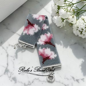 Op de afbeelding: Een kralenarmband met een bloemmotief in roze en wit op een grijze achtergrond. De armband heeft een zilveren sluiting en is voorzien van het label "Ralu's Bead Art". De armband is ongeveer 18 cm lang.