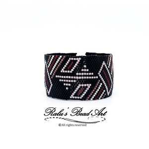 Può includere: Braccialetto nero con perline con un motivo geometrico in tonalità rosa, bianco e rosso. Il braccialetto è realizzato con perline a seme e presenta un design intrecciato.