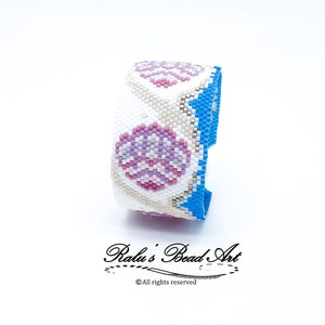 Op de afbeelding: Een witte kralen armband met een blauwe rand en een roze en wit geometrisch patroon. De armband is gemaakt met kleine kraaltjes en heeft een delicate, vrouwelijke uitstraling.