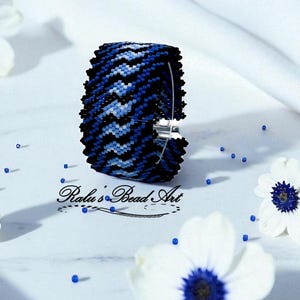 Könnte beinhalten: Ein Armreif aus blauen und schwarzen Perlen mit Zickzackmuster. Der Armreif hat einen silbernen Verschluss und ist von weißen Blumen mit blauen Zentren umgeben. Der Text "Ralu's Bead Art" ist sichtbar.