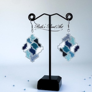 May include: A pair of handmade beaded earrings in a geometric design. The earrings feature a mosaic pattern of white, light blue, teal, and navy blue beads. The earrings are displayed on a black stand against a white background.