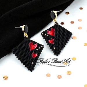 Puede incluir: Pendientes negros con forma de diamante y cuentas con detalles de corazones rojos. Los pendientes tienen postes dorados y tapas circulares negras. El texto "Ralu's Bead Art" es visible.
