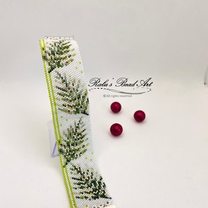 May include: A white beaded bracelet with a green leaf pattern. The bracelet has a silver clasp and is displayed on a white surface with red beads.