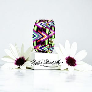 May include: A wide, colorful beaded bracelet with a geometric pattern in black, purple, green, orange, and white. The bracelet is displayed on a white block with the text "Ralu's Bead Art". Two white flowers with purple centers flank the bracelet.