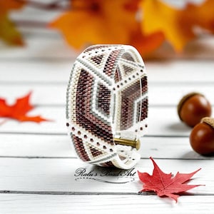 May include: A beaded cuff bracelet with geometric patterns in shades of white, brown, and gray. The bracelet has a gold-colored clasp. The background includes fall leaves and acorns.