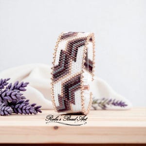 May include: A beaded cuff bracelet with a geometric pattern in white, brown, gray, and gold. The bracelet is approximately 1.5 inches wide and features a copper clasp. The background includes lavender sprigs and the text "Ralu's Bead Art".