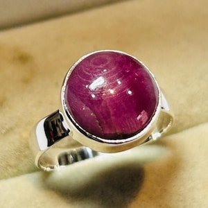 May include: A silver ring featuring a large, round, polished, deep red gemstone. The gemstone is set in a simple, modern silver band. The ring is displayed in a beige jewelry box, highlighting the ring's color and design.