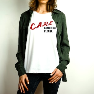 May include: White t-shirt with the text "C.A.R.E. ABOUT ME PLEASE." in red and black. The shirt is worn under a dark green button-up shirt and paired with dark jeans. The image is set against a white background.