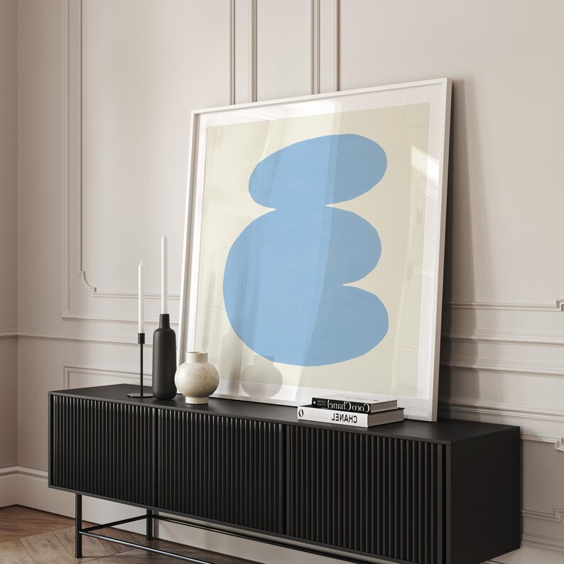 May include: A framed abstract art print featuring a blue, organic shape against a cream background. The print is displayed on a black media console with two books on top.