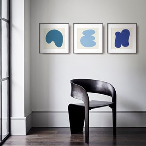 May include: Three framed abstract art prints featuring blue and white shapes on a white background. The prints are hanging on a white wall above a black wooden chair.