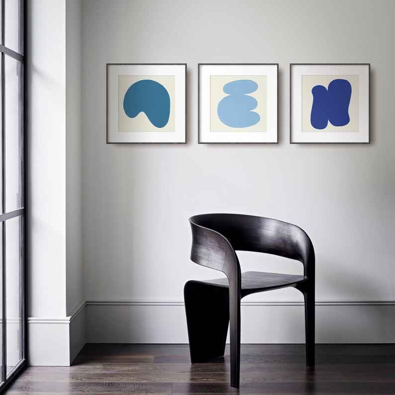 May include: Three framed abstract art prints featuring blue and white shapes on a white background. The prints are hanging on a white wall above a black wooden chair.
