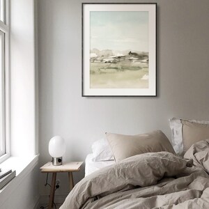 Op de afbeelding: Een ingelijst aquarel schilderij van een landschap met een lichtblauwe lucht, groene heuvels en een bruin veld. Het schilderij hangt aan een lichtgrijze muur boven een bed met een beige dekbed en kussens.