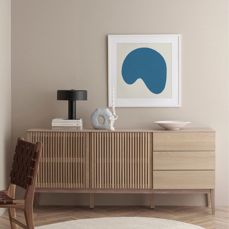 May include: A light wood cabinet with two doors and three drawers. The cabinet has a slatted design on the doors. A black lamp with a white lampshade sits on top of the cabinet. A white ceramic vase with a hole in the side sits on the cabinet. A white bowl sits on the cabinet. A framed print with a blue abstract shape hangs on the wall above the cabinet.