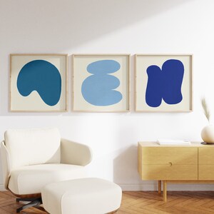 May include: Three framed abstract art prints featuring blue and cream shapes. The prints are hanging on a white wall above a light wood dresser.