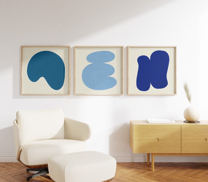May include: Three framed abstract art prints featuring blue and cream shapes. The prints are hanging on a white wall above a light wood dresser.