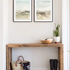 Op de afbeelding: Een rustieke houten consoletafel met een geweven mand, een paar zwarte regenlaarzen, een plant in een pot en een houten kom erop. Twee ingelijste aquarel landschapsschilderijen hangen aan de muur boven de tafel.