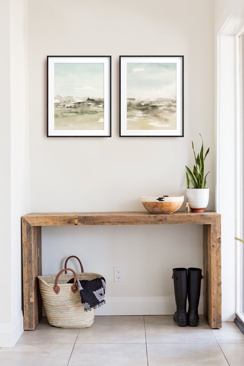 Op de afbeelding: Een rustieke houten consoletafel met een geweven mand, een paar zwarte regenlaarzen, een plant in een pot en een houten kom erop. Twee ingelijste aquarel landschapsschilderijen hangen aan de muur boven de tafel.