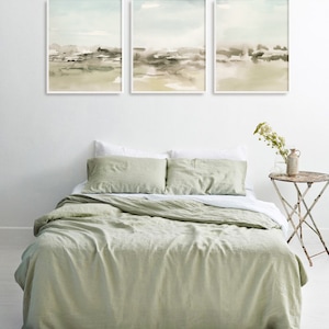 Op de afbeelding: Drie ingelijste aquarellen van abstracte landschappen in tinten groen, bruin en wit hangen boven een dekbedovertrek van saliegroen linnen op een bed met witte kussenslopen.