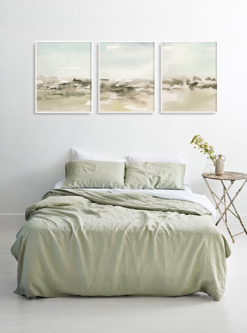 Op de afbeelding: Drie ingelijste aquarellen van abstracte landschappen in tinten groen, bruin en wit hangen boven een dekbedovertrek van saliegroen linnen op een bed met witte kussenslopen.
