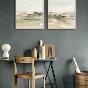 Op de afbeelding: Twee ingelijste aquarellen van abstracte landschappen in tinten groen, bruin en beige, hangend aan een grijze muur boven een houten bureau met een stoel, een lamp en andere decoratie.