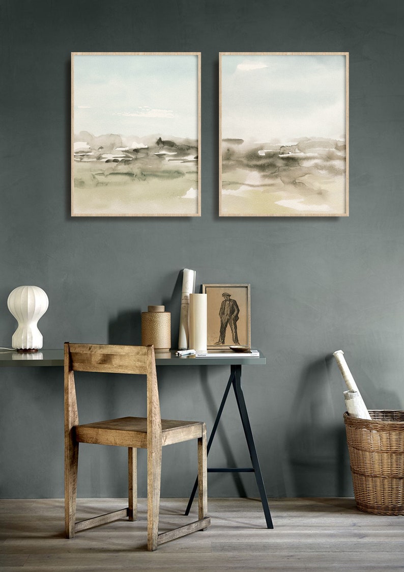 Op de afbeelding: Twee ingelijste aquarellen van abstracte landschappen in tinten groen, bruin en beige, hangend aan een grijze muur boven een houten bureau met een stoel, een lamp en andere decoratie.