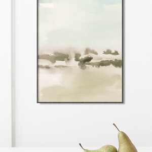 Op de afbeelding: Een ingelijst abstract aquarel schilderij met een lichtblauw, beige en bruin kleurenschema. Het schilderij toont een landschap met een wazige, atmosferische sfeer.