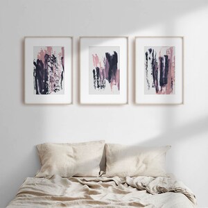 May include: Three framed abstract paintings with pink and purple brushstrokes on a white background. The paintings are hanging on a white wall above a beige linen bed.