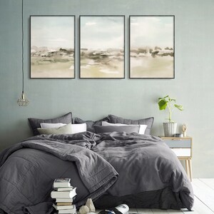 Op de afbeelding: Drie ingelijste abstracte aquarellen in gedempte groene en bruine tinten, hangend aan een lichtblauwe muur boven een grijs bed met een grijs dekbed en witte kussens.